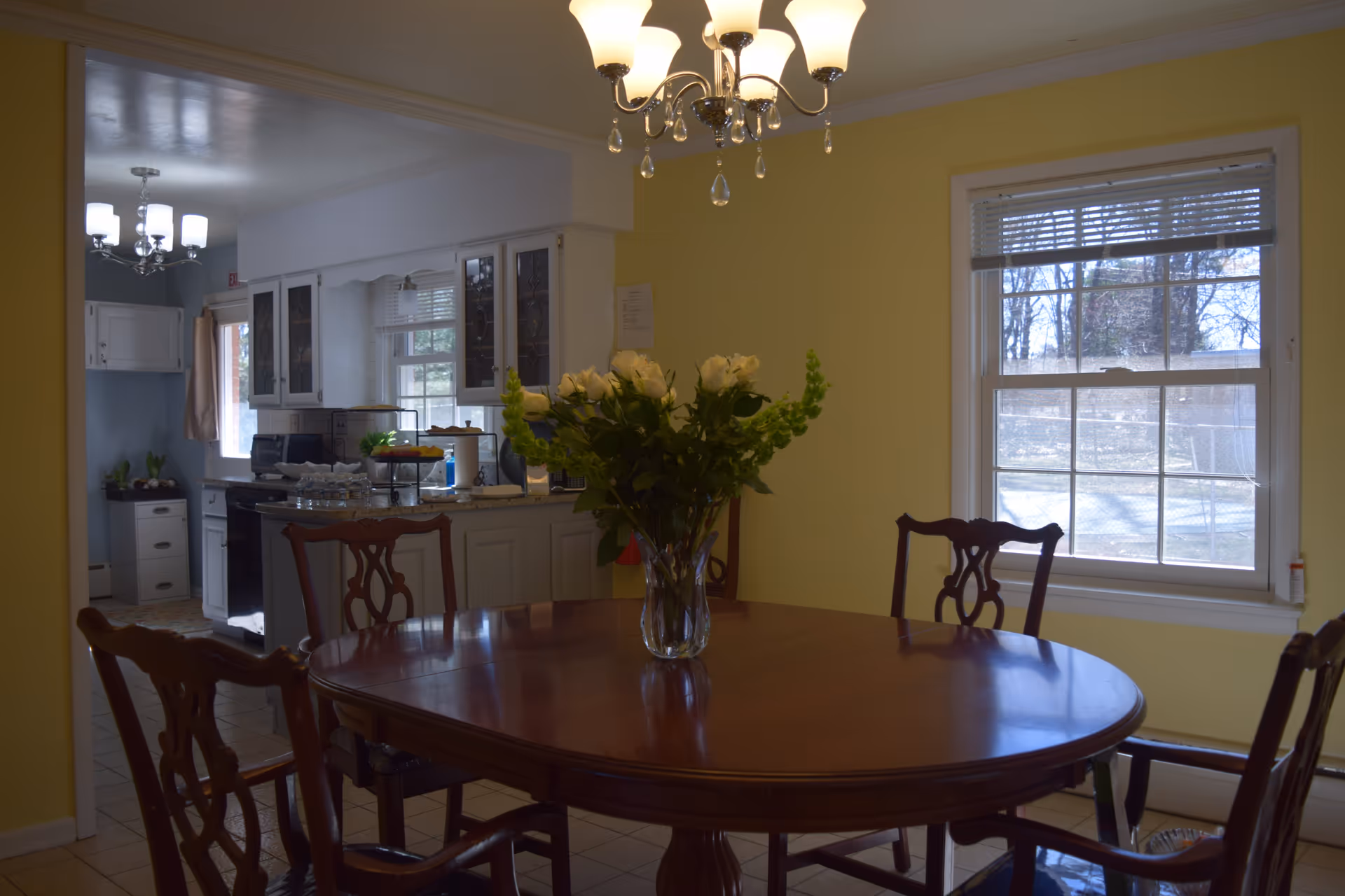 Dining room with a wooden table and chairs, a vase of flowers in the center, and a view into an adjacent kitchen.