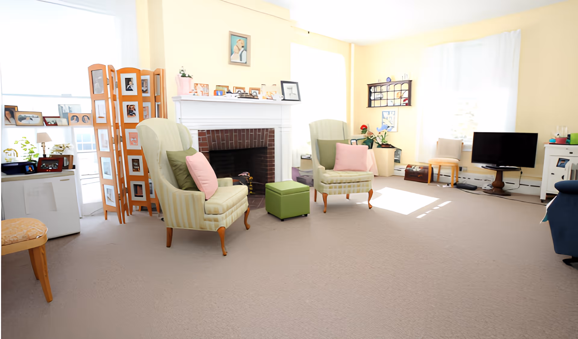 A bright living room with two striped armchairs each with green and pink cushions, a green ottoman, a white fireplace with framed photos on the mantel, a wooden folding screen with photos, a TV on a small wooden table, and large windows with white curtains letting in natural light.