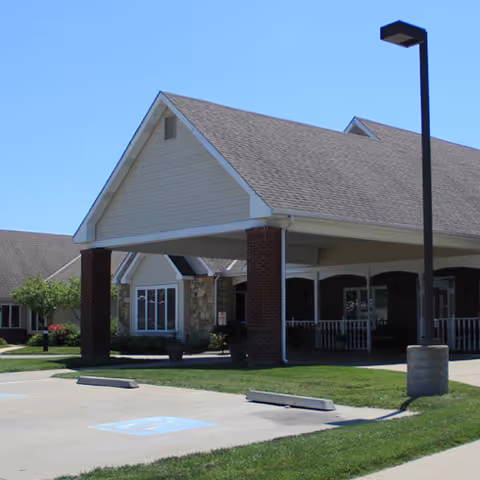 Exterior view of a senior living facility building with a covered entrance supported by brick columns, a parking lot with a handicapped parking space, and a streetlamp on the right side.