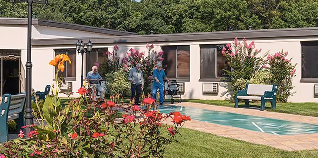 Outdoor courtyard area at The Arbors facility with three elderly people walking along a paved path. The courtyard features green grass, blooming flowers, benches, and a shuffleboard court. The building with windows and flowering bushes is visible in the background.