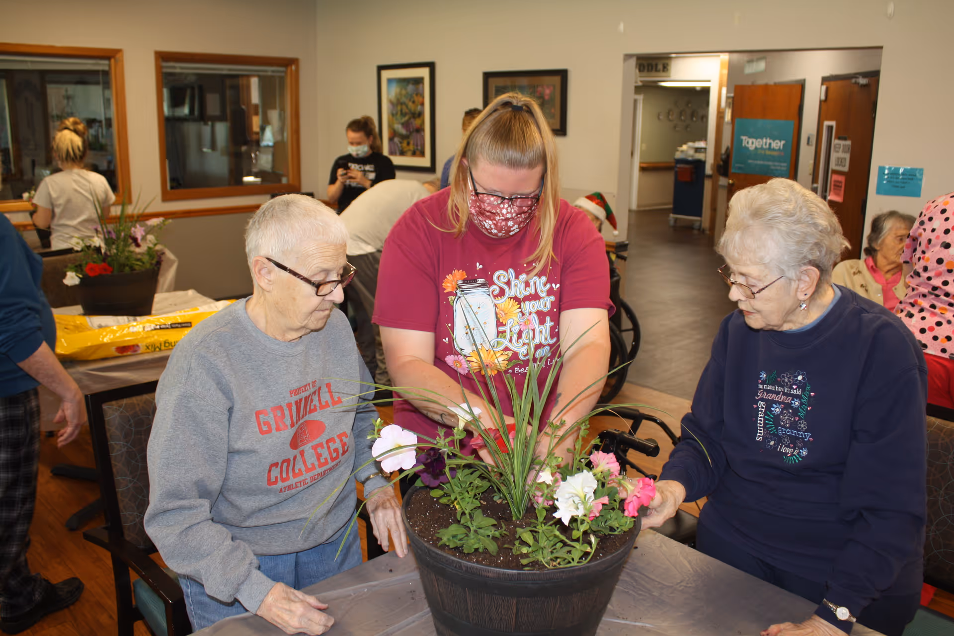 Three women, two elderly and one younger wearing a mask, are sitting around a table indoors working on a flower planting activity with a large pot of colorful flowers in front of them. Other people are visible in the background in a communal area with light-colored walls and wooden floors.