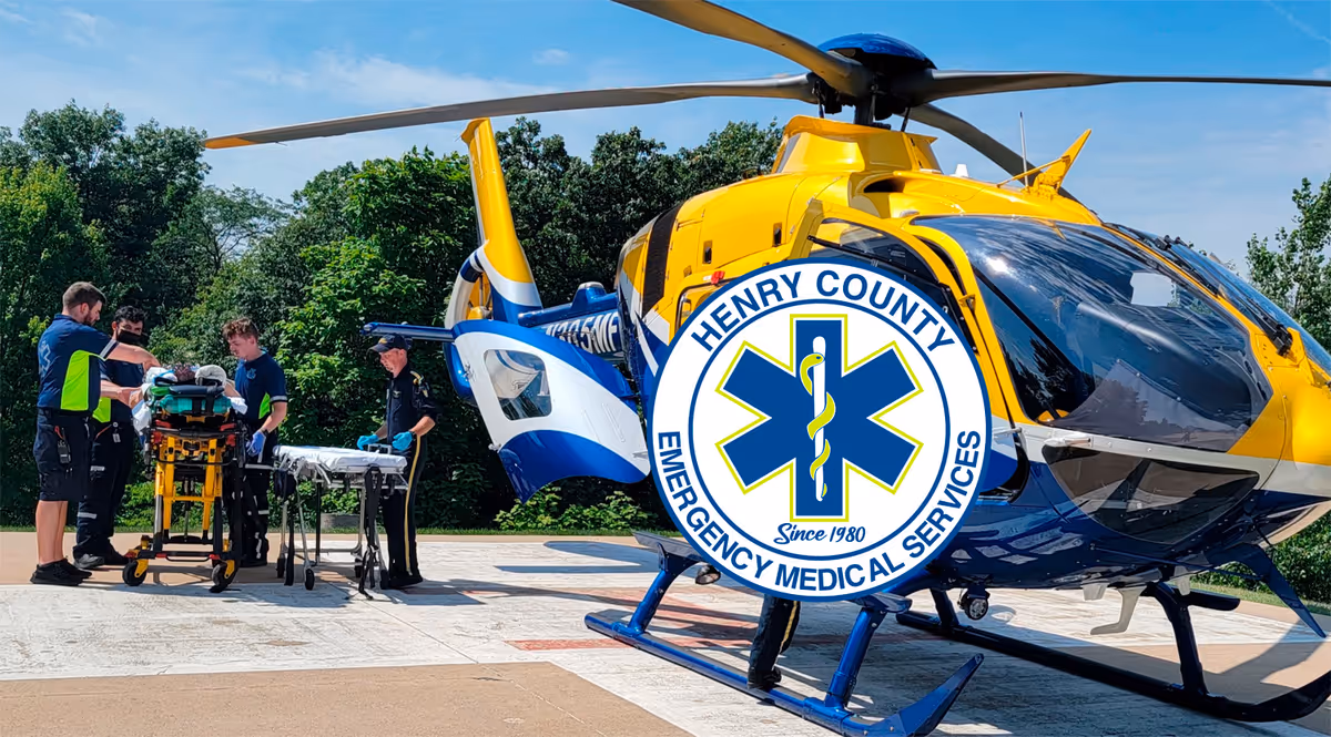 A yellow-and-blue medical helicopter on a helipad with emergency personnel loading a stretcher nearby.