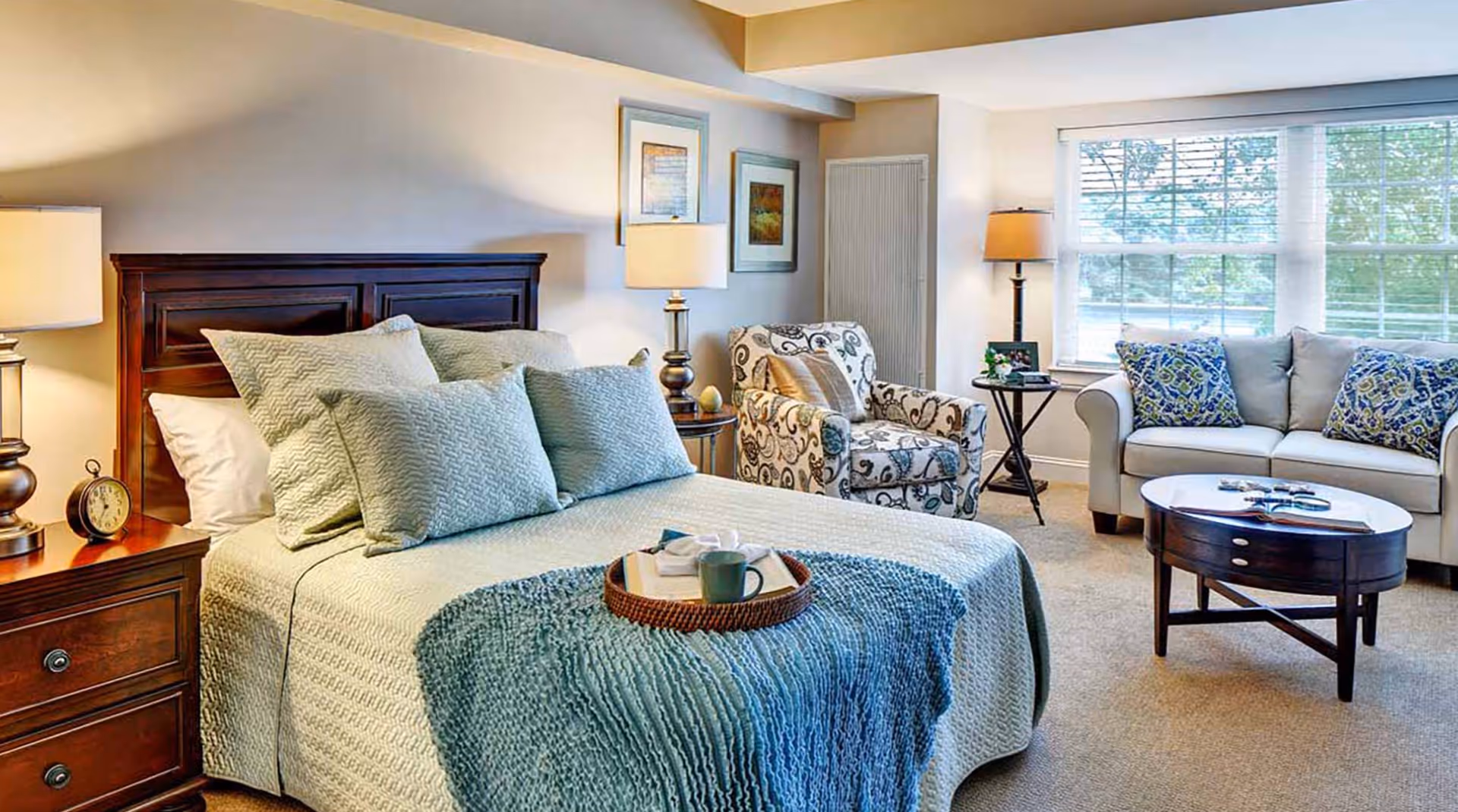 A cozy bedroom with a neatly made bed featuring multiple pillows and a blue throw blanket. There is a wooden nightstand with a lamp and an alarm clock on the left side of the bed. To the right, there is a patterned armchair, a small side table with a lamp, and a beige sofa with decorative pillows near a large window letting in natural light. A round wooden coffee table is placed in front of the sofa.