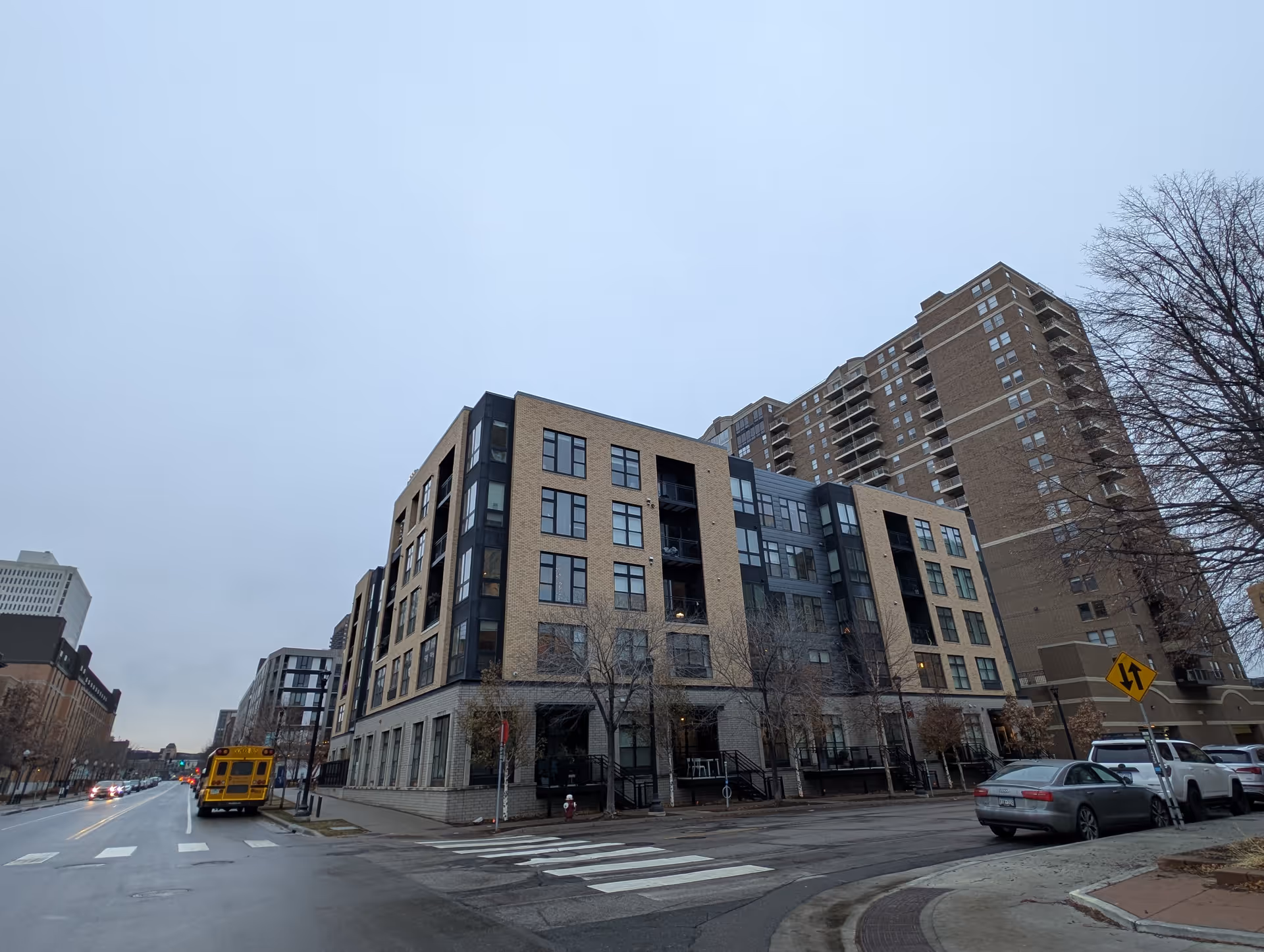 Street view of a multi-story residential building on a cloudy day, with parked cars and a yellow school bus on the road nearby. Leafless trees line the sidewalk.