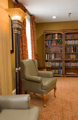 A cozy interior room featuring two green leather armchairs, a tall decorative floor lamp, patterned carpet, and a wooden bookshelf filled with books and decorative items. The room has warm yellow walls and red patterned curtains covering a window.