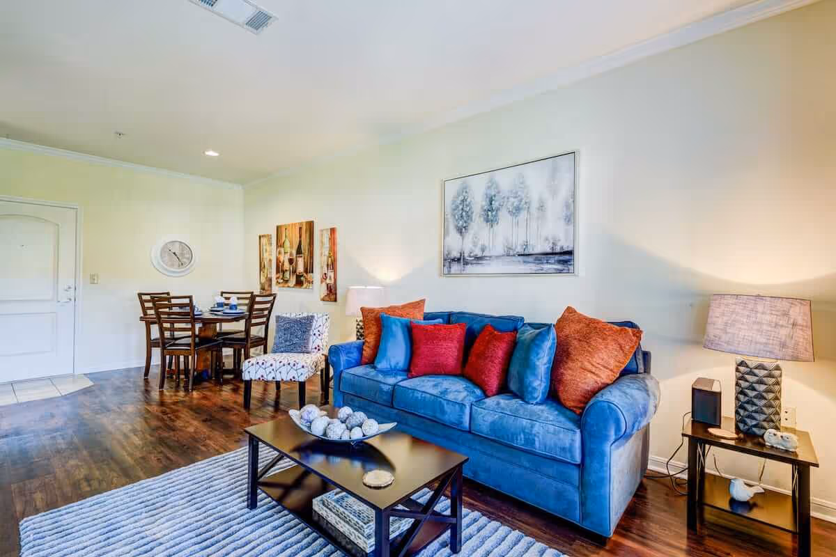 A cozy living room area with a blue sofa adorned with red and blue cushions, a black coffee table with decorative items, a side table with a lamp, and a patterned armchair. In the background, there is a small dining table with four chairs, wall art, and a clock on a light-colored wall. The floor is wooden with a light-colored rug in front of the sofa.