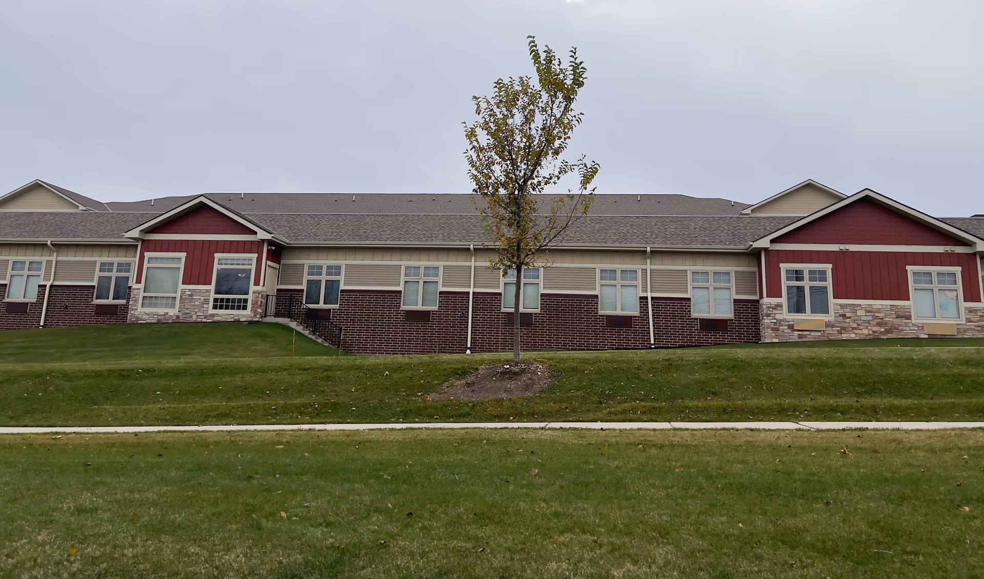 Exterior view of a single-story building with a combination of red, beige, and stone siding. There is a small tree planted on a grassy lawn in front of the building, with a sidewalk running parallel to the building.