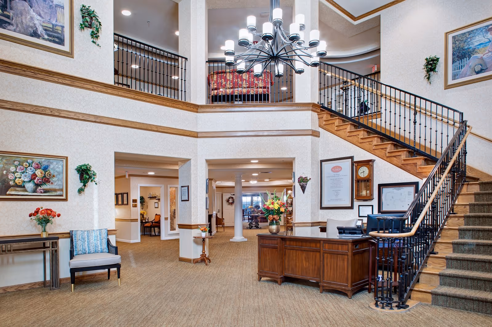 Spacious and well-lit senior living facility lobby with a wooden reception desk, a staircase with black iron railings leading to an upper floor, floral paintings on the walls, a chandelier hanging from the ceiling, and comfortable seating areas.