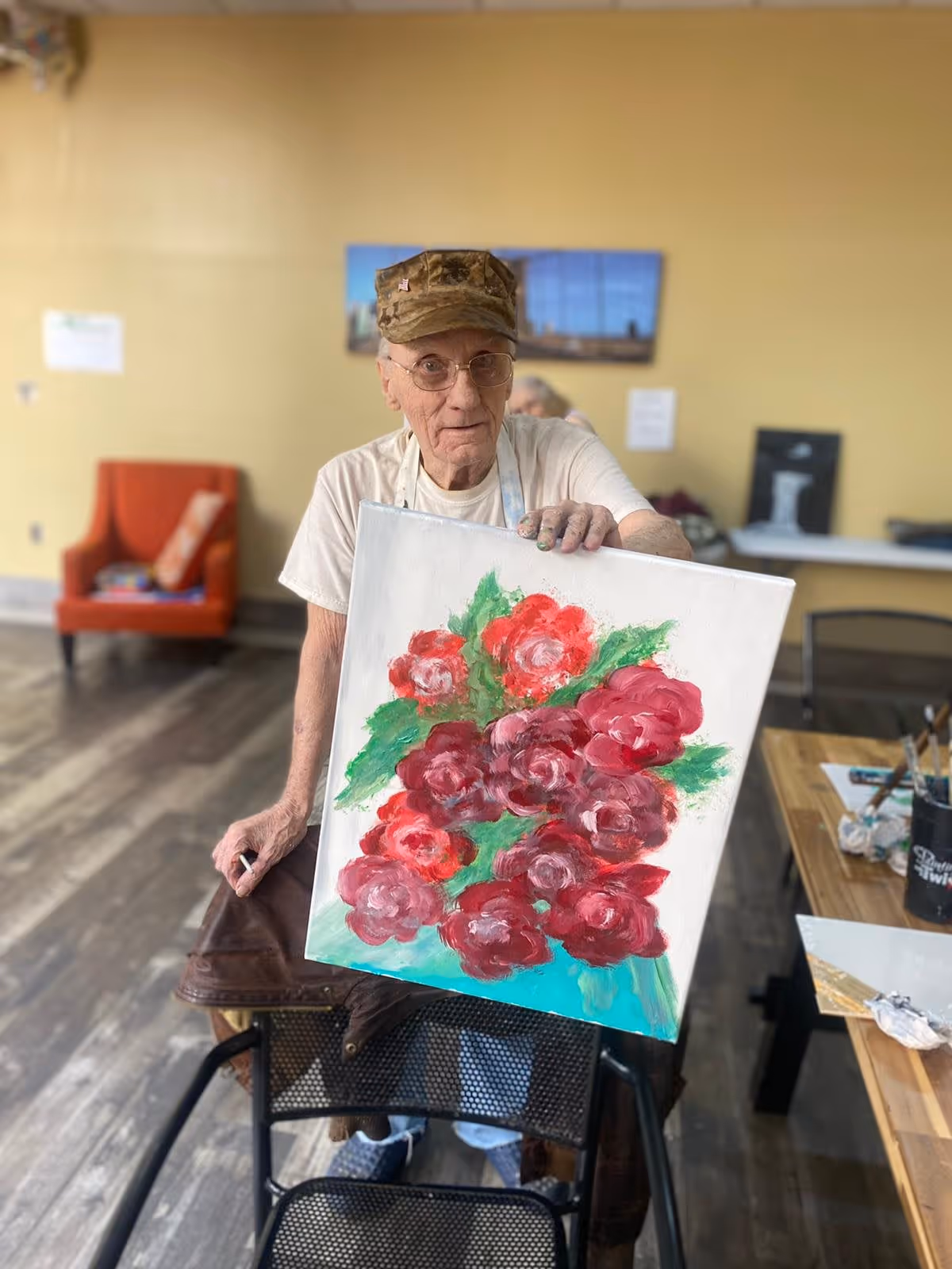 An elderly man wearing a camouflage cap and glasses is sitting indoors, holding up a canvas painting of red and pink flowers with green leaves. The room has wooden flooring, a yellow wall, an orange armchair, and a table with painting supplies.