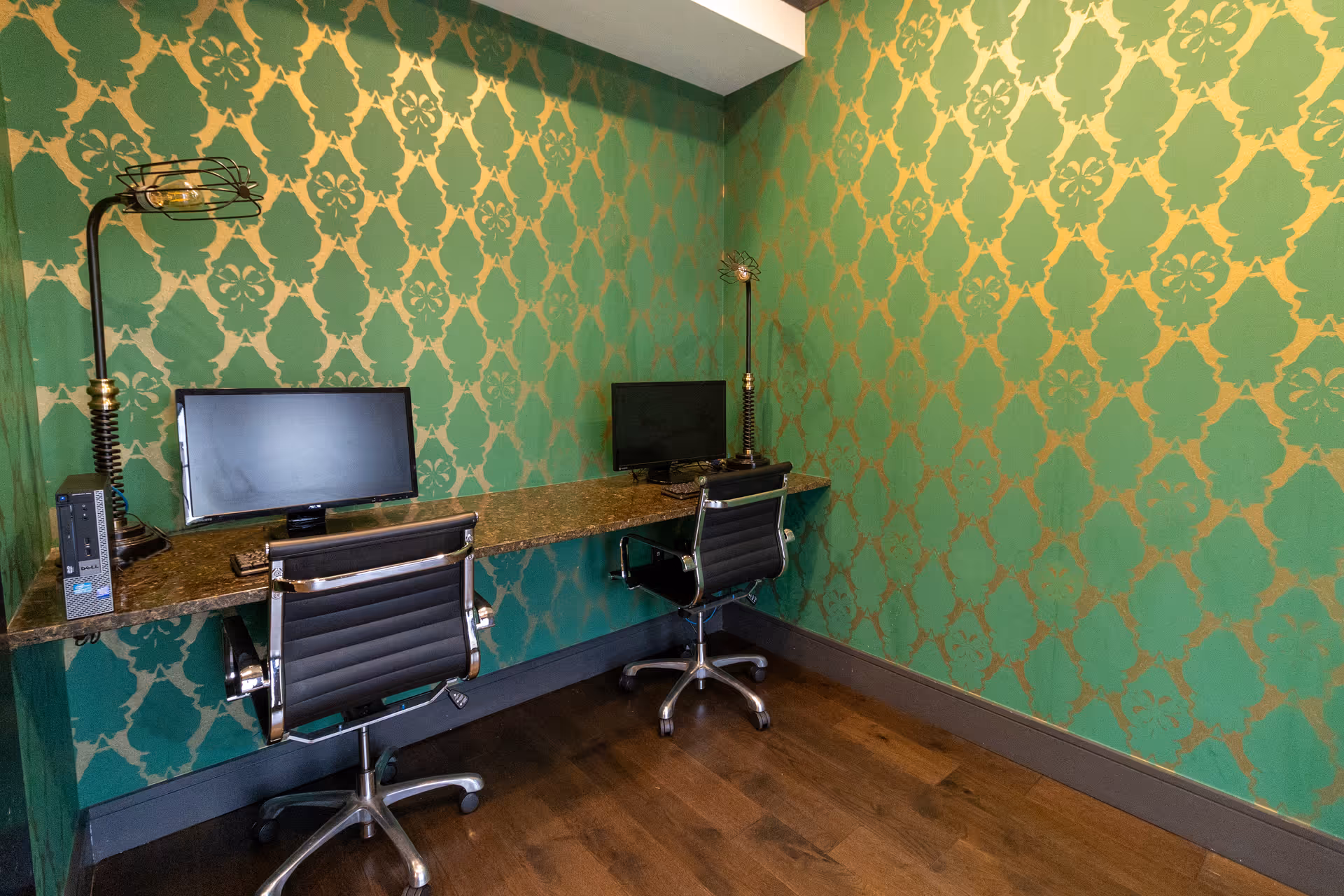 A small office or computer room with two black office chairs in front of a long granite desk. Two computer monitors and keyboards are placed on the desk, along with two decorative lamps. The walls are covered with green and gold patterned wallpaper, and the floor is wooden.