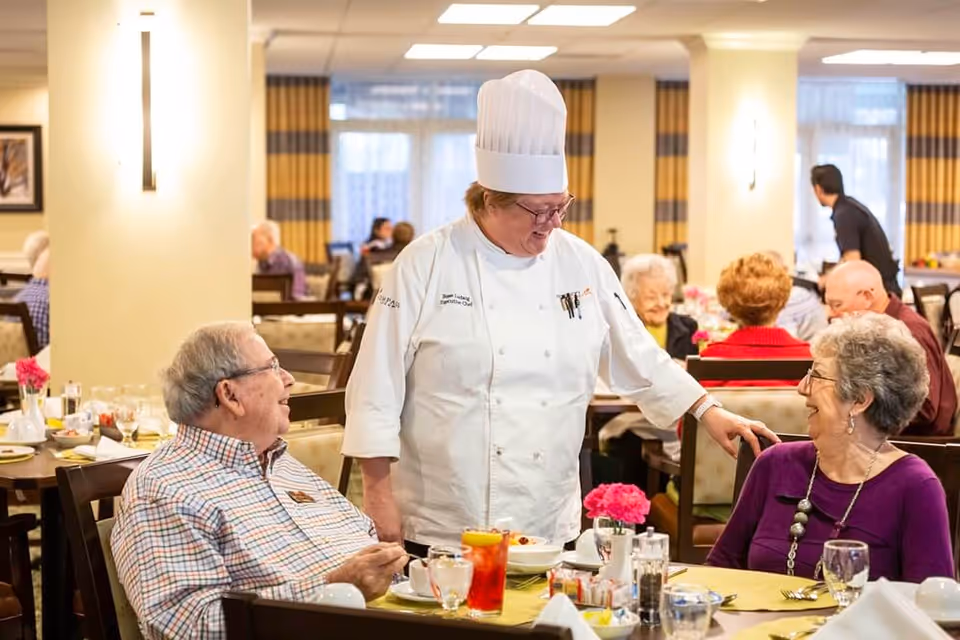 A chef in a white uniform and tall chef hat smiling and interacting with two elderly people seated at a dining table in a well-lit dining room. The table is set with glasses, plates, and a red drink, and there are pink flowers as a centerpiece. Other elderly people are visible in the background, also seated and dining.