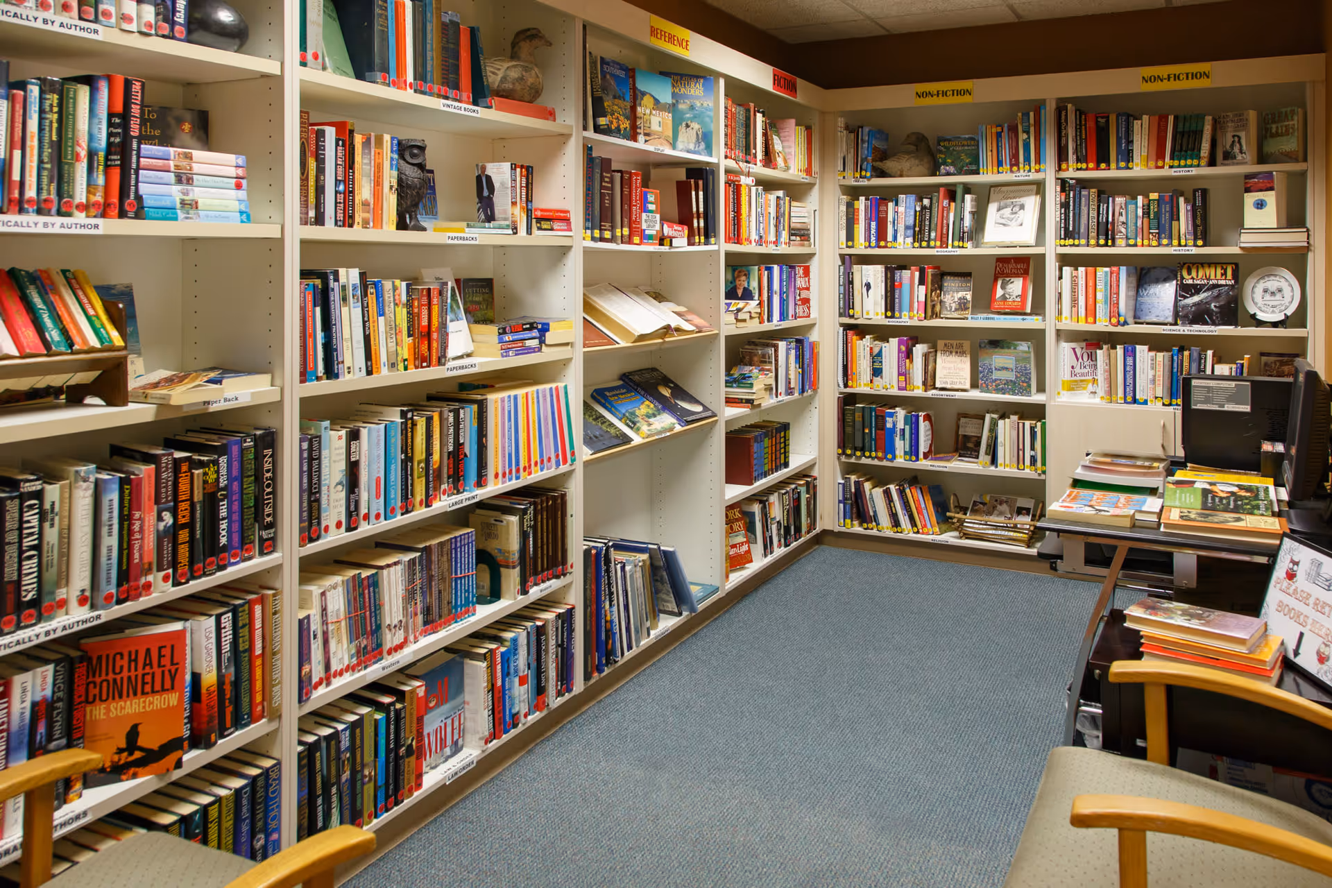 Small interior library with wall-to-wall bookshelves, chairs, a desk and a computer.