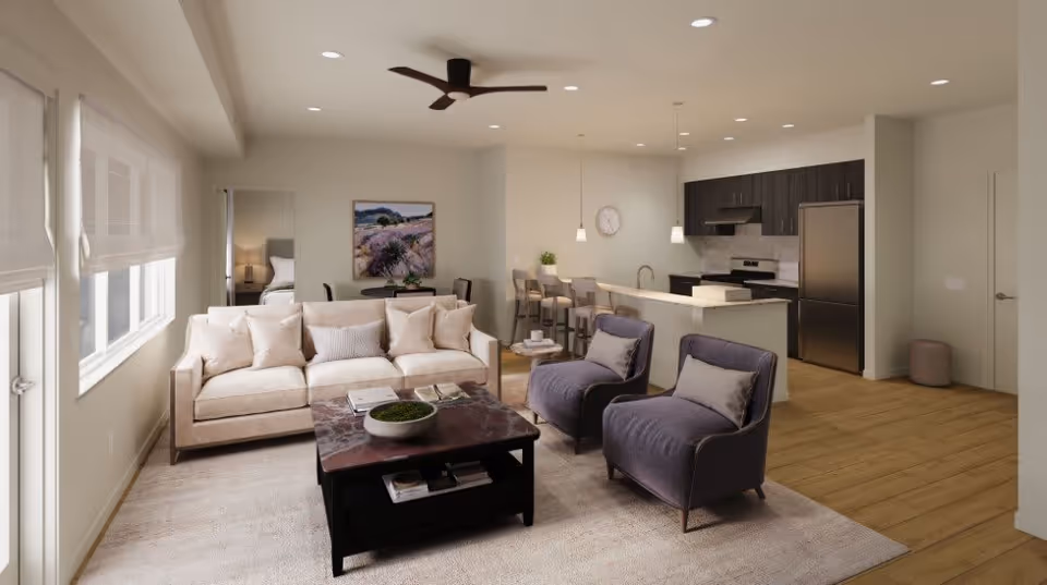 A modern senior living apartment interior featuring a beige sofa with multiple cushions, two purple armchairs, a dark wooden coffee table with decorative items, and a light-colored rug. The open floor plan includes a kitchen area with a breakfast bar, three bar stools, stainless steel refrigerator, stove, and pendant lighting. A ceiling fan is mounted above, and a bedroom is partially visible through an open doorway. The walls are painted light beige, and large windows with white blinds allow natural light to fill the space.