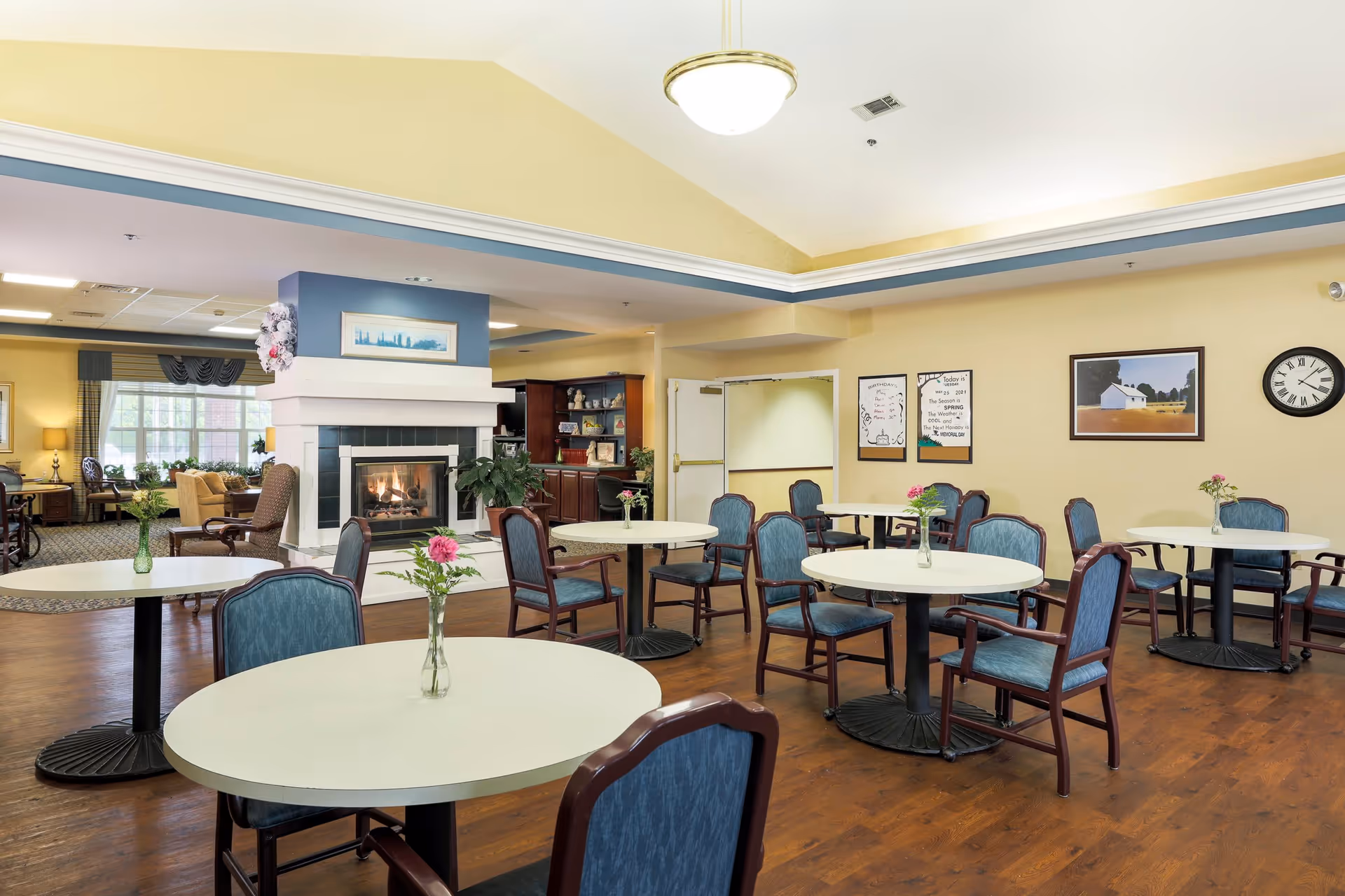 A spacious dining area in an assisted living facility with round tables and blue cushioned chairs. Each table has a small vase with flowers. There is a fireplace with a lit fire in the background, along with a seating area and large windows letting in natural light. The walls are painted yellow and decorated with framed pictures and a clock.