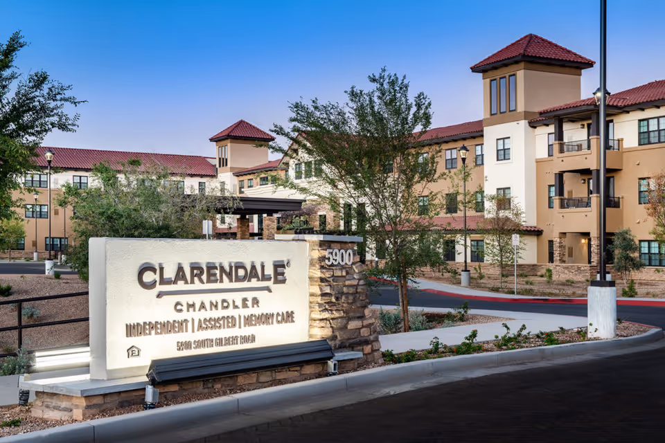 Exterior view of Clarendale Of Chandler senior living facility at dusk, showing a large sign with the facility name and services offered, surrounded by trees and landscaped areas, with the multi-story building in the background featuring balconies and a red-tiled roof.