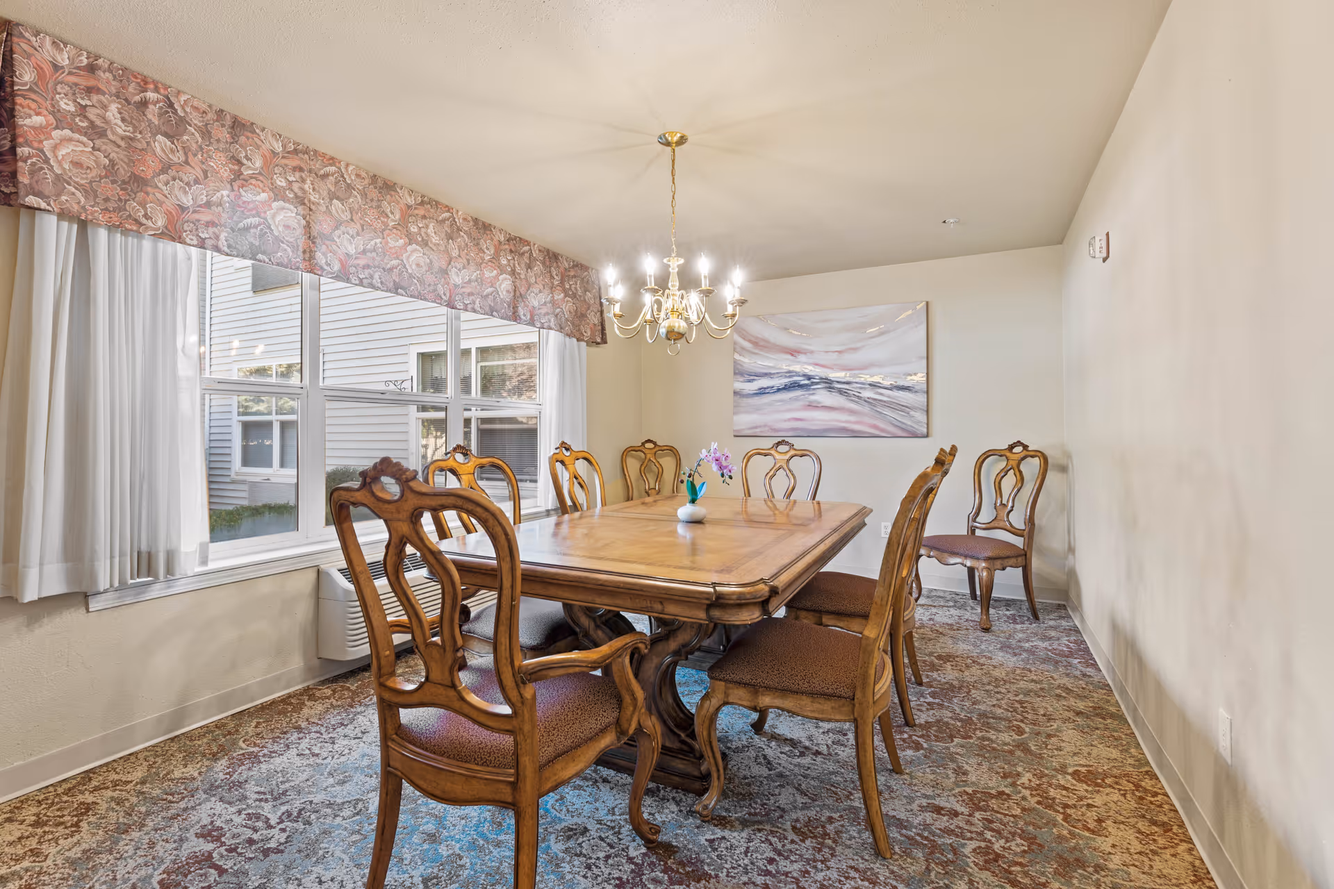 A dining room with a wooden table surrounded by eight wooden chairs with cushioned seats. A chandelier with lit bulbs hangs above the table. There is a large window with floral valance and white curtains on the left wall, and a framed abstract painting on the far wall. The floor is covered with a patterned carpet.