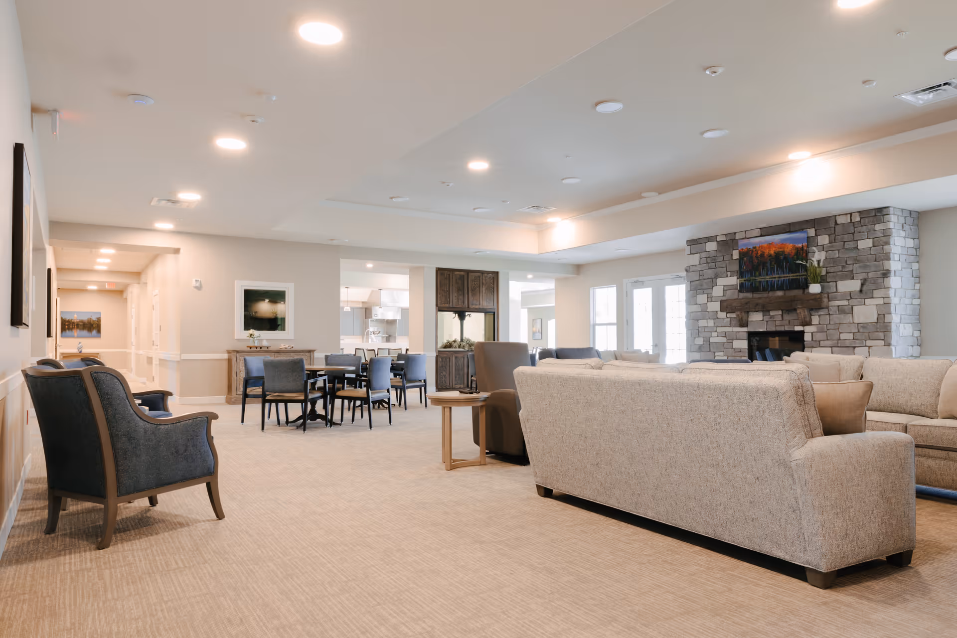 A spacious and well-lit living room area in a senior living facility featuring beige sofas, a stone fireplace with a mounted TV above it, several chairs, and a dining area with tables and chairs in the background.