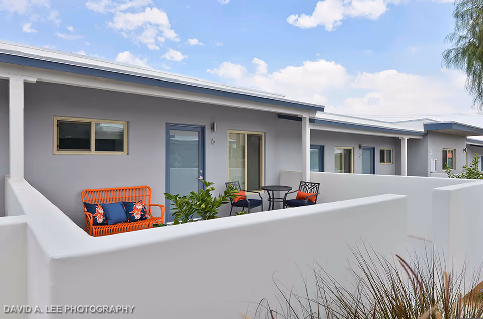 Outdoor patio area of Stonewall Gardens Assisted Living with white walls, a bright orange bench with blue and floral cushions, two black chairs with orange cushions, a small round table, and some green plants. The building exterior is light gray with blue doors and windows under a partly cloudy sky.