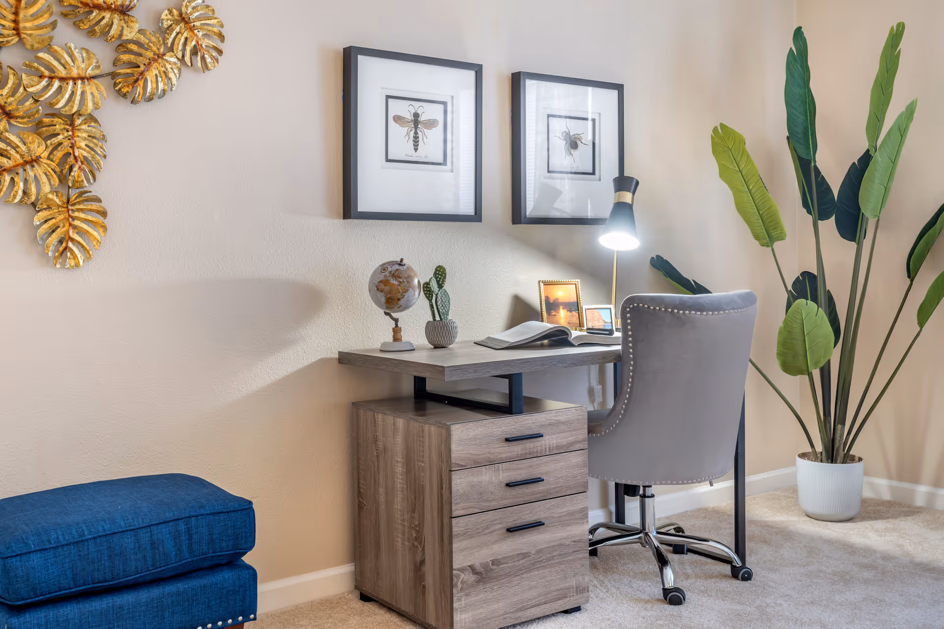 A cozy home office setup featuring a wooden desk with three drawers, a gray upholstered swivel chair, a desk lamp, a small globe, a potted cactus, and an open book. Two framed insect illustrations hang on the beige wall above the desk. To the right, there is a tall green potted plant, and to the left, part of a blue cushioned ottoman is visible. A decorative golden leaf wall art is mounted on the left side of the wall.