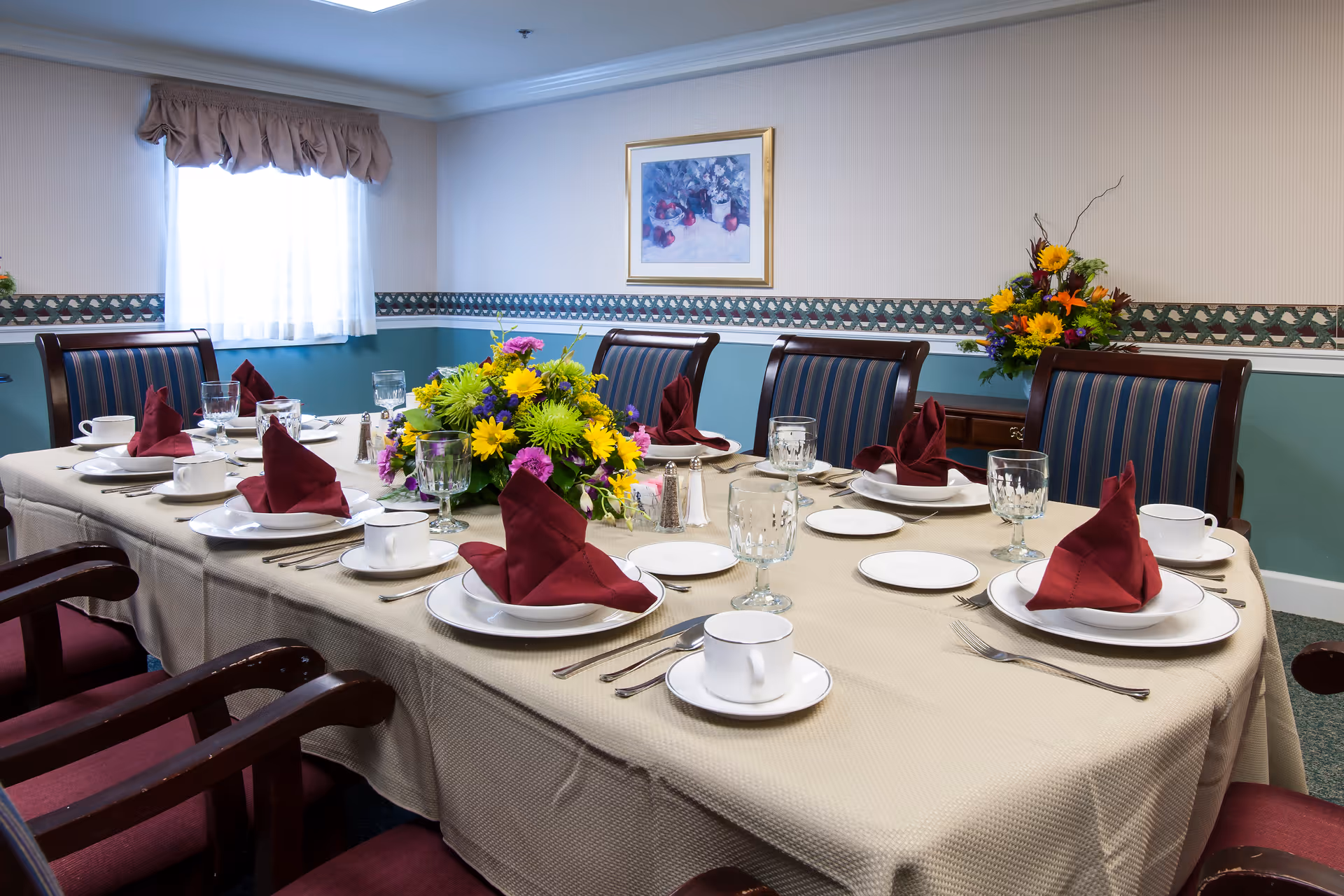 A dining room table set for eight with beige tablecloth, white plates, cups, silverware, crystal glasses, and burgundy folded napkins. A colorful floral centerpiece is in the middle of the table. The room has teal and beige walls with a decorative border, a window with a valance, a framed painting, and a floral arrangement on a side table.