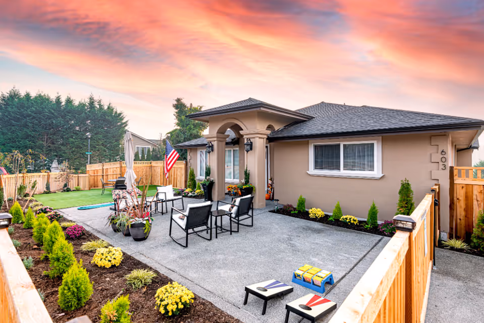 Outdoor patio area of a single-story residential building with a seating arrangement of four chairs and a small table. The patio is surrounded by a wooden fence and landscaped garden beds with small shrubs and yellow flowers. An American flag is displayed near the entrance, and the sky shows a colorful sunset with pink and orange hues.