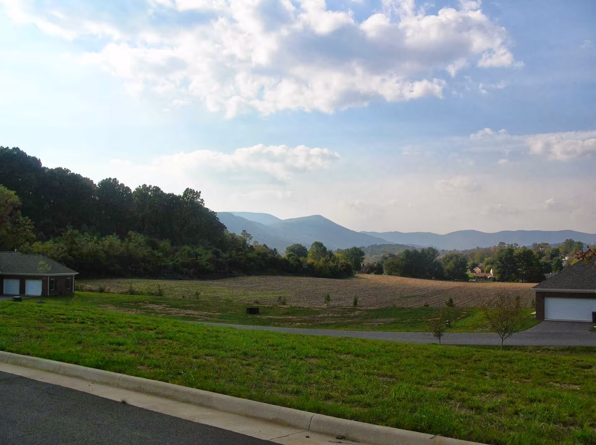 A scenic outdoor view showing a grassy area with a paved road in the foreground, two small buildings with garages on either side, and a large open field with trees and hills in the background under a partly cloudy sky.