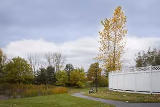 A winding paved pathway through a grassy area with trees showing autumn foliage under a cloudy sky. A white fence curves along the right side of the path.