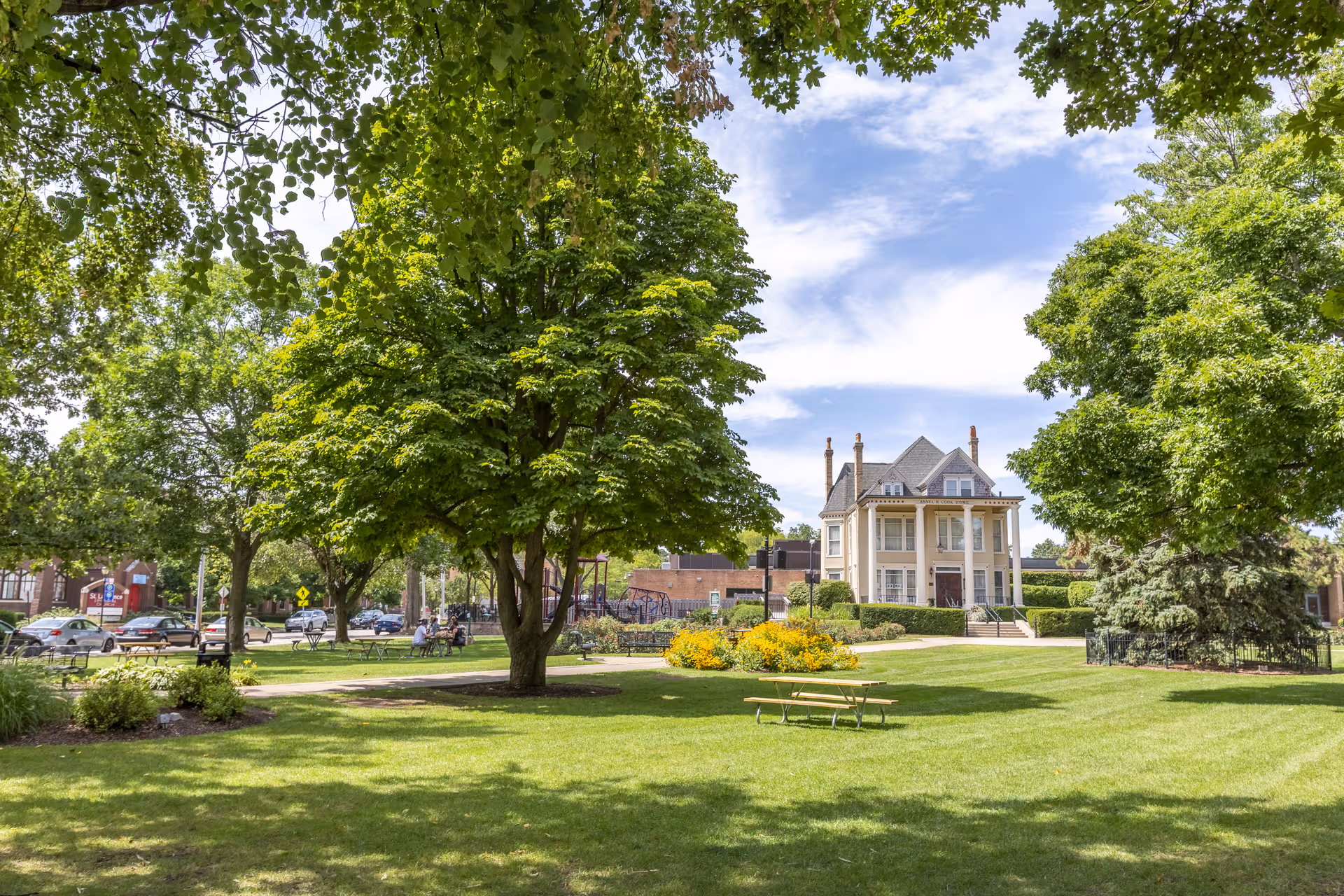 A sunny outdoor scene featuring a well-maintained green lawn with large leafy trees and a picnic table. In the background, there is a large, elegant two-story house with a porch and multiple chimneys. Several cars are parked along the street, and a few people are sitting on benches near a playground area.
