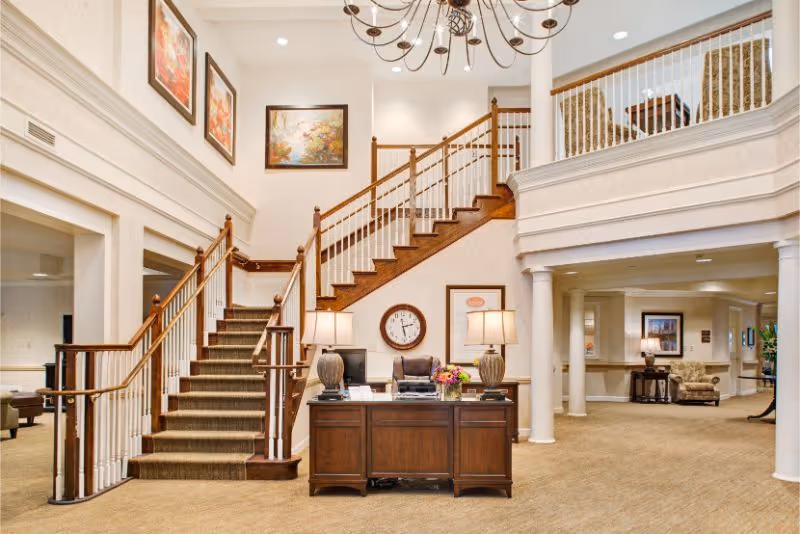 Spacious reception lobby with a central wooden desk, twin staircases leading to an upper balcony, lamps, and framed artwork.