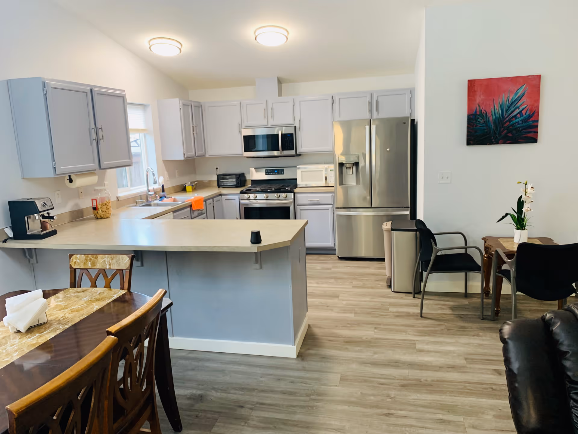 A modern kitchen and dining area with light gray cabinets, stainless steel appliances including a refrigerator, microwave, and stove. There is a kitchen island with a beige countertop, a dining table with wooden chairs, and a small seating area with two black chairs and a small wooden table with a white orchid plant. The floor is light wood, and a colorful painting with red and green tones hangs on the wall.