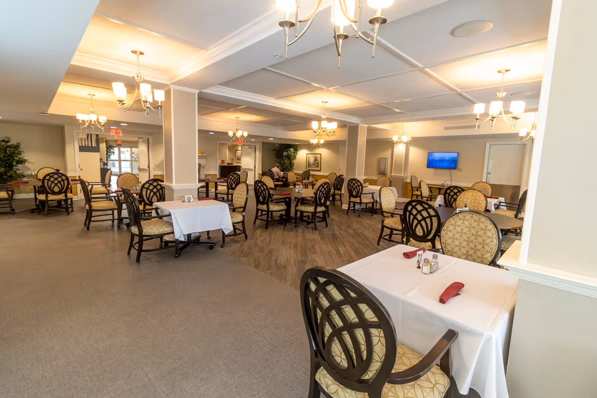 Spacious senior living dining room with multiple set tables and patterned cushioned chairs under chandeliers.