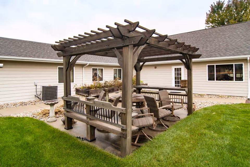 Outdoor seating area with a wooden pergola covering a round table surrounded by cushioned chairs and benches, situated on a concrete patio next to a building with beige siding and windows, with green grass and landscaping rocks around.