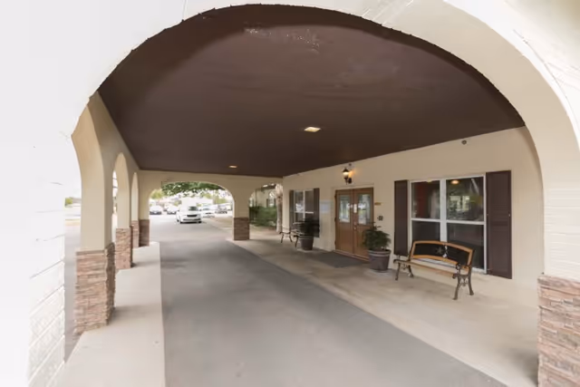 Covered entrance area of a building with arched openings, benches, potted plants, and a double-door entrance. Cars are visible in the background outside the covered area.