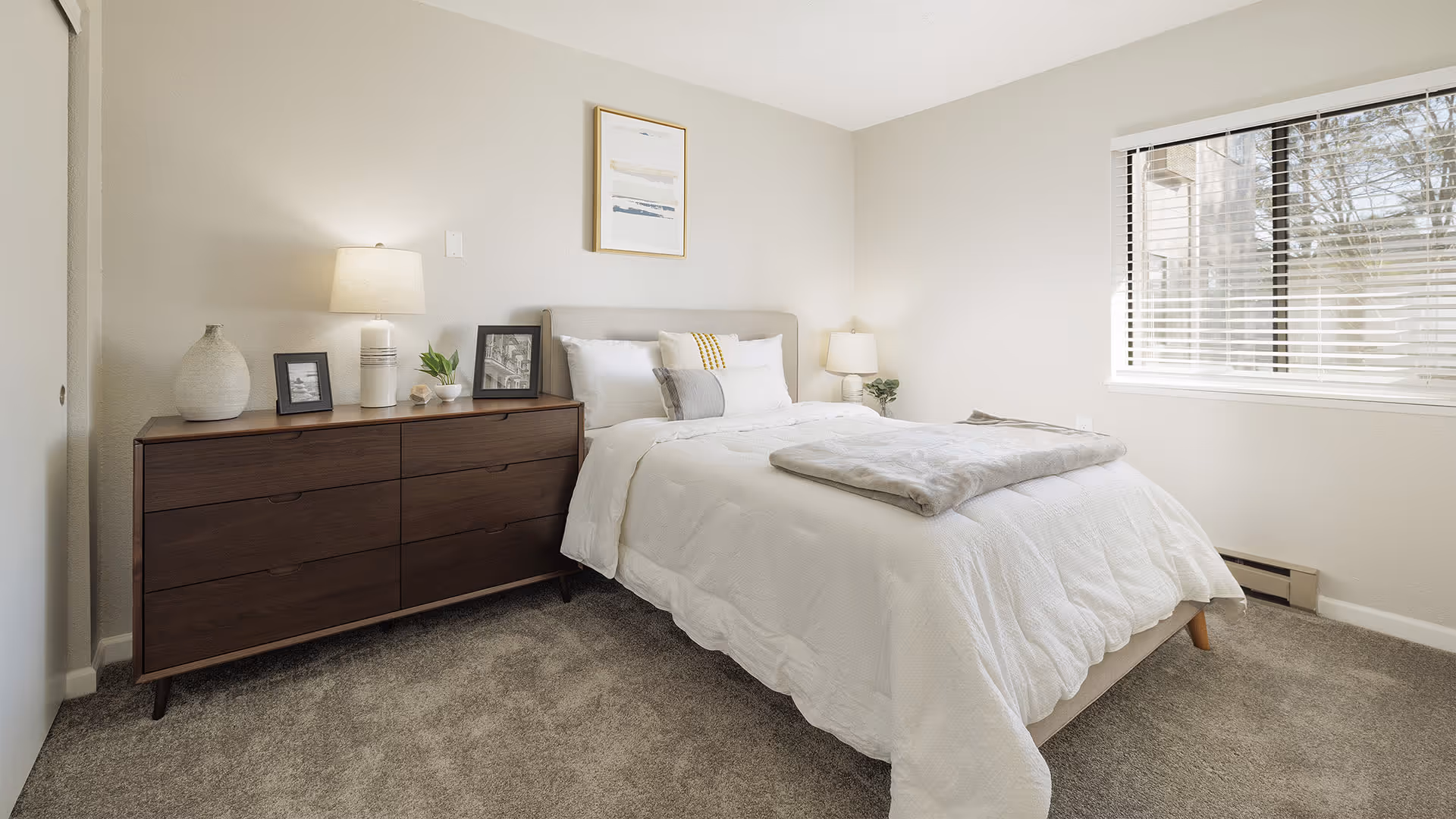 A bright and clean bedroom with a large window on the right letting in natural light. The room features a neatly made bed with white bedding and a gray throw blanket at the foot. To the left of the bed is a dark wooden dresser with a lamp, framed photos, a vase, and a small plant. The walls are painted light beige, and there is a framed abstract artwork hanging above the bed.
