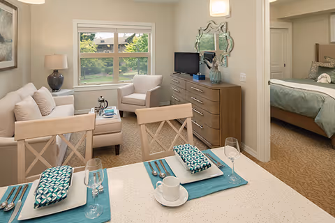 A cozy assisted living apartment featuring a small dining area with two place settings, a living room with a sofa, armchair, coffee table, and a lamp, and a bedroom visible through an open doorway. The room has neutral tones with teal accents and a window showing greenery outside.