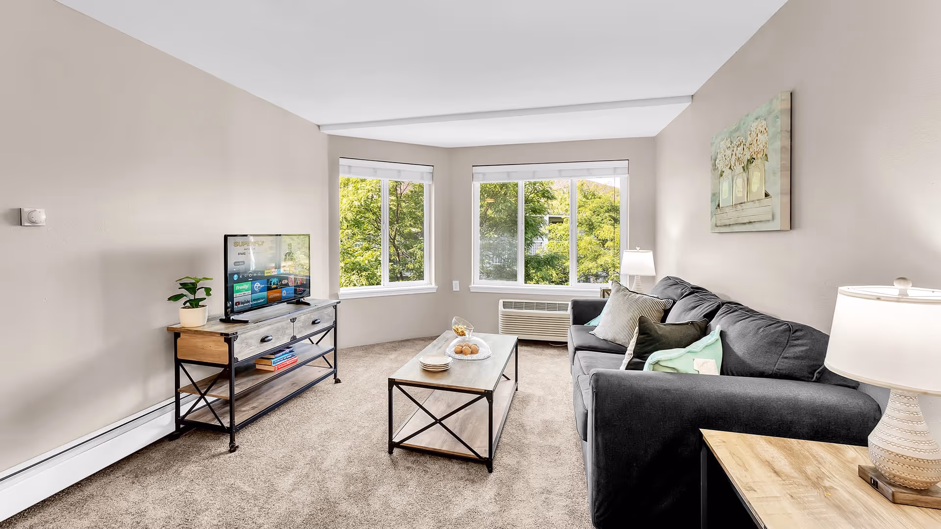 A bright living room with beige walls and carpeted floor featuring a dark gray sofa with multiple cushions, a wooden coffee table with plates and a glass bowl, a TV on a wooden and metal stand, two large windows with views of green trees, and two table lamps providing soft lighting.