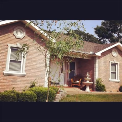 Exterior view of a single-story brick house with white trim around windows and roof edges. There is a small porch with a chair and a decorative water fountain. A young tree and some bushes are planted in the front yard with a grassy lawn.
