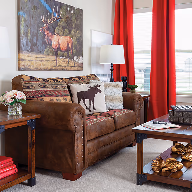Cozy living room with a brown leather couch adorned with patterned cushions, including one with a moose silhouette. A painting of a moose hangs on the wall above the couch. There is a wooden side table with a vase of flowers and red books, and a wooden coffee table with decorative golden fish, an open book, and glasses. Red curtains frame a window letting in natural light.