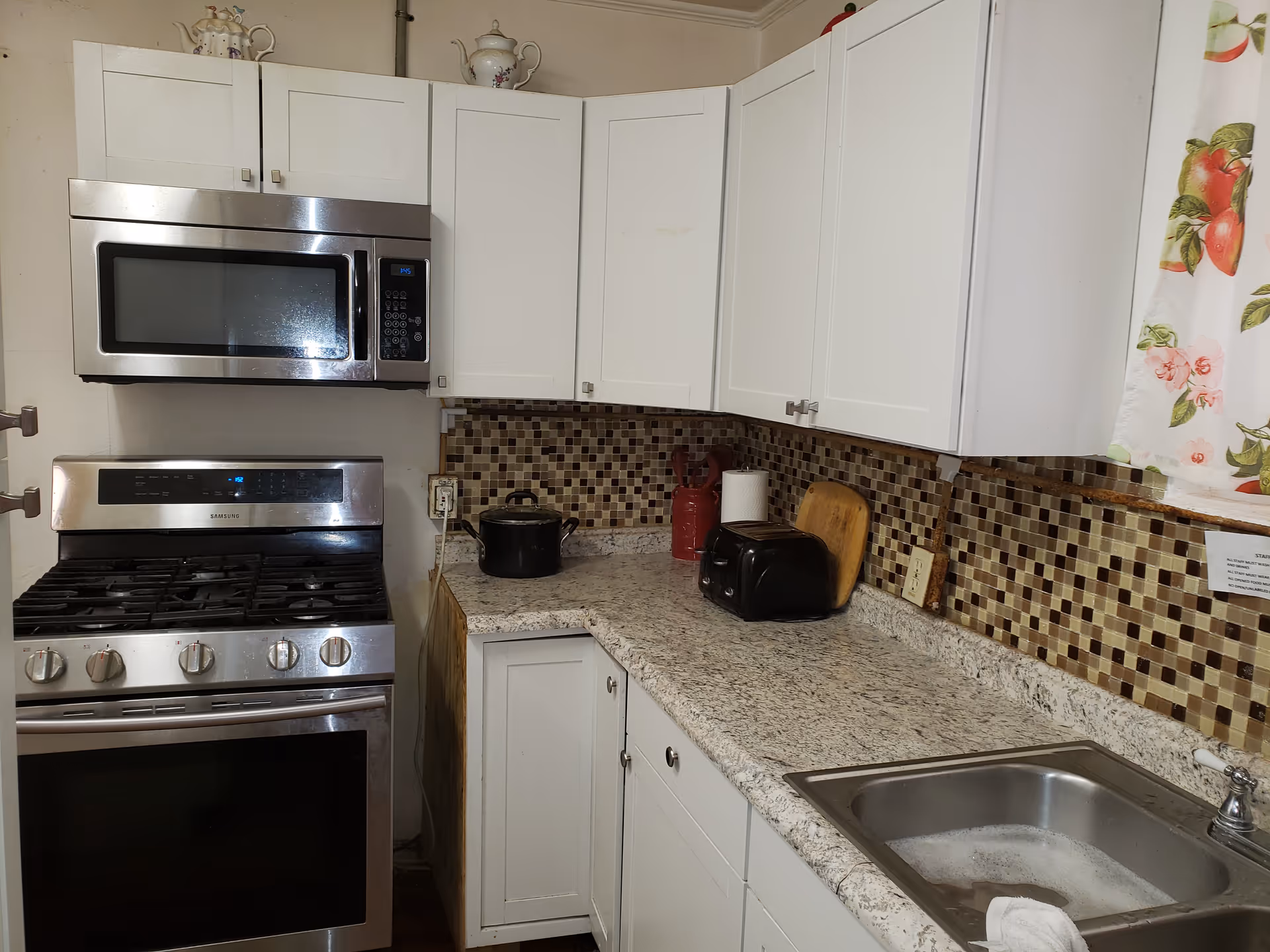 A kitchen area with white cabinets, a stainless steel microwave mounted above a stainless steel gas stove, a granite countertop with a black pot, a red container, a paper towel holder, a black toaster, a wooden cutting board, and a stainless steel sink filled with soapy water. The backsplash features small square mosaic tiles in shades of brown and beige. A floral curtain is partially visible on the right side.