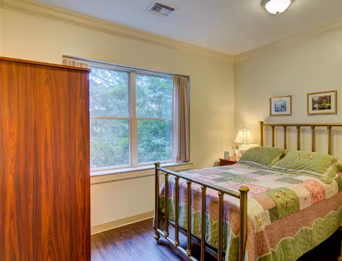 A cozy bedroom in Rosecrest Retirement Community featuring a brass bed with a patchwork quilt, a wooden wardrobe, two framed pictures on the wall, a bedside table with a lamp, and a large window with beige curtains overlooking greenery outside.