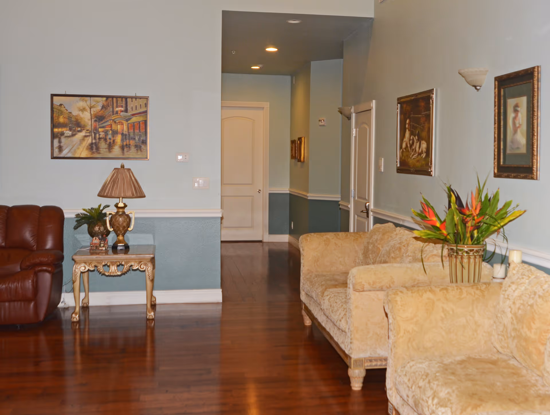 Interior view of a living room area in an assisted living facility with wooden flooring, a brown leather armchair, a glass-top side table with a decorative lamp and plant, beige upholstered sofas, framed paintings on light blue walls, and a hallway leading to closed doors.