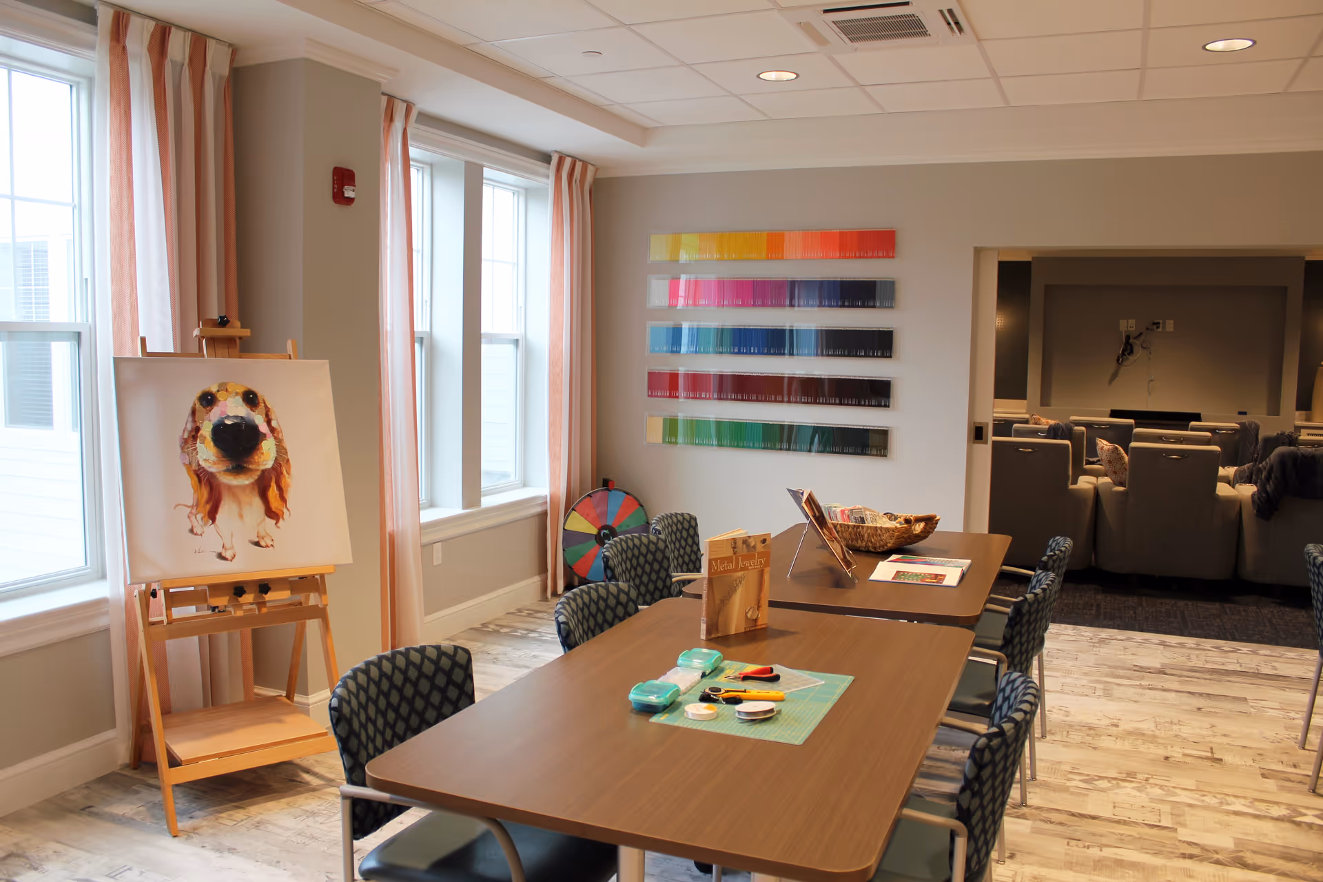 Bright interior activity room with a long table and chairs, an easel displaying a dog painting, colorful wall swatches, and a seating area in the background.