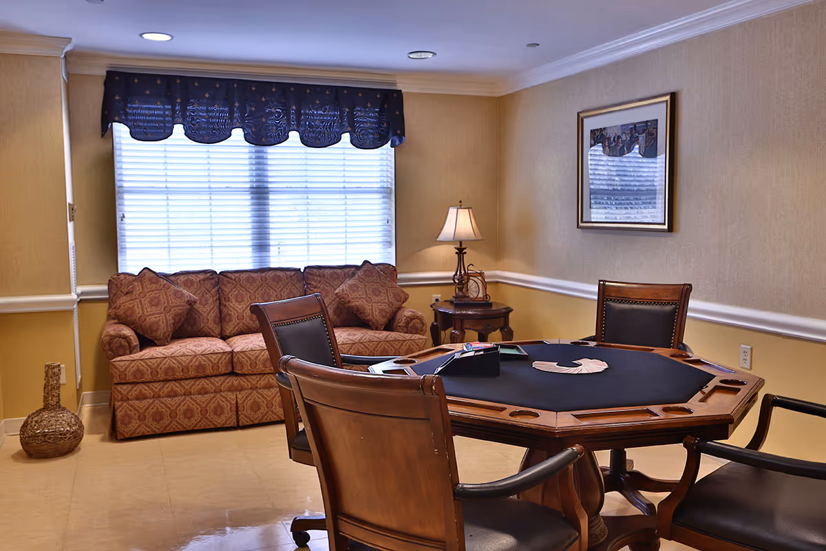A cozy room with a patterned sofa against a window with blinds and a dark valance. In front of the sofa is a wooden octagonal card table with a black felt top, surrounded by four wooden chairs with black leather seats. A small round side table with a lamp is placed in the corner, and a framed picture hangs on the wall.