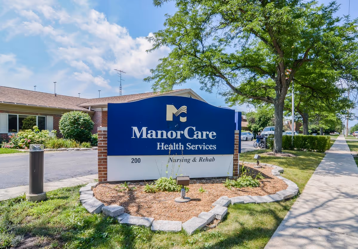 Outdoor view of a ManorCare Health Services sign for Nursing & Rehab, situated in a landscaped area with mulch and plants. Behind the sign is a single-story brick building with windows and shrubs. A sidewalk and trees line the street on the right side under a partly cloudy sky.
