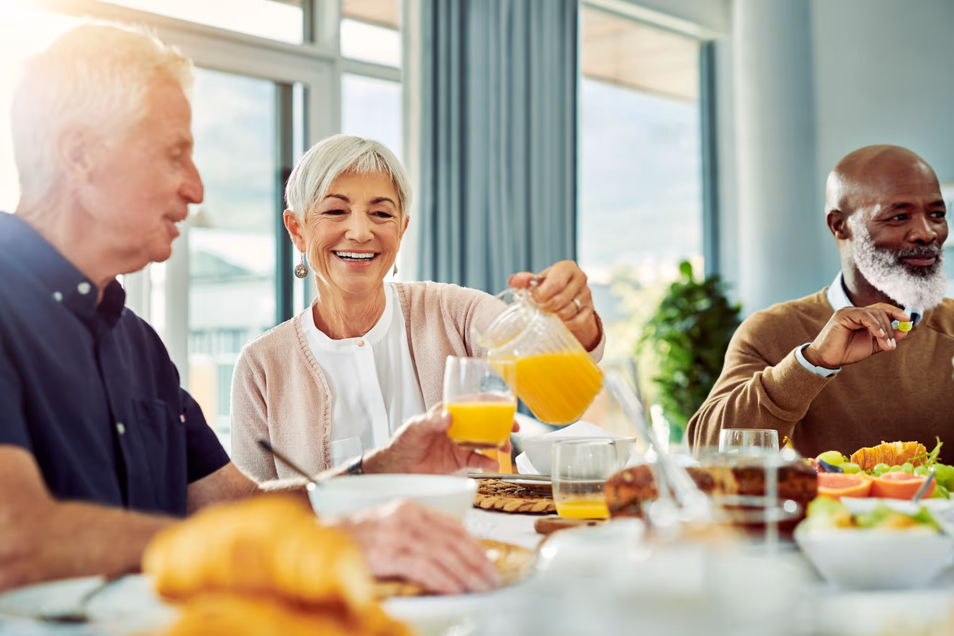 Three older adults sit around a dining table as a woman pours orange juice into a glass.