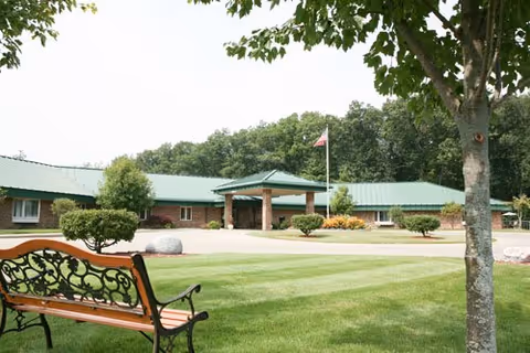 View of a single-story senior living facility building with a green roof and brick exterior, surrounded by well-maintained green lawns, trees, and shrubs. A wooden bench with decorative metal armrests is in the foreground, and an American flag flies near the building entrance.