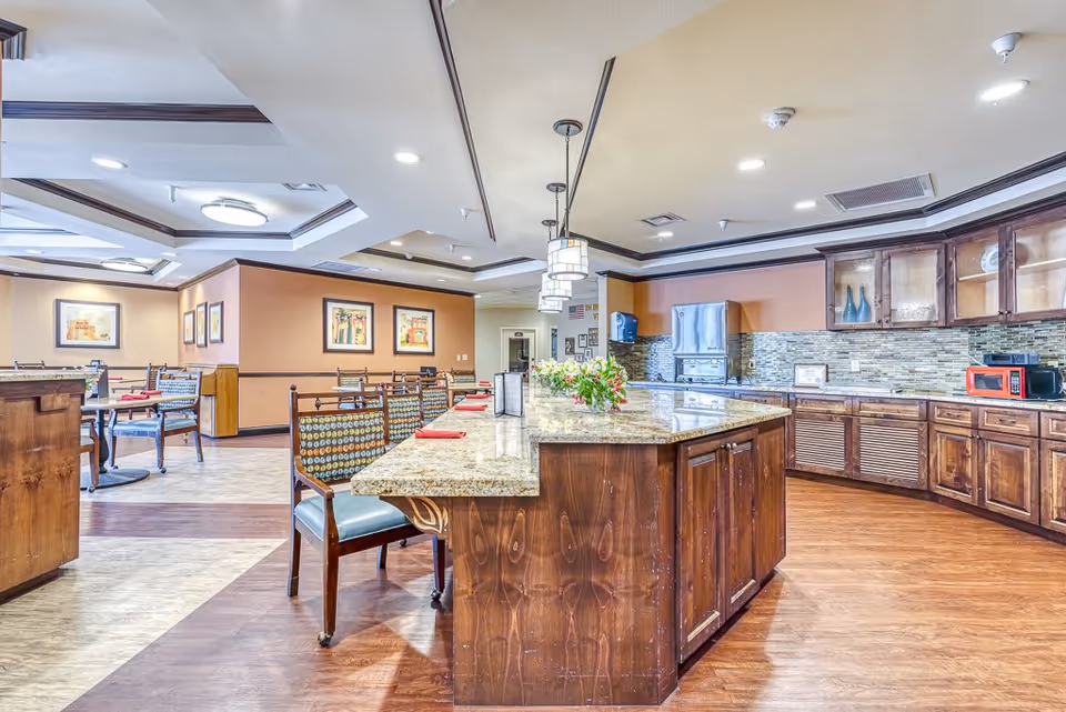 Bright communal dining area with a central granite-topped island, wooden cabinetry, and multiple dining tables and chairs.