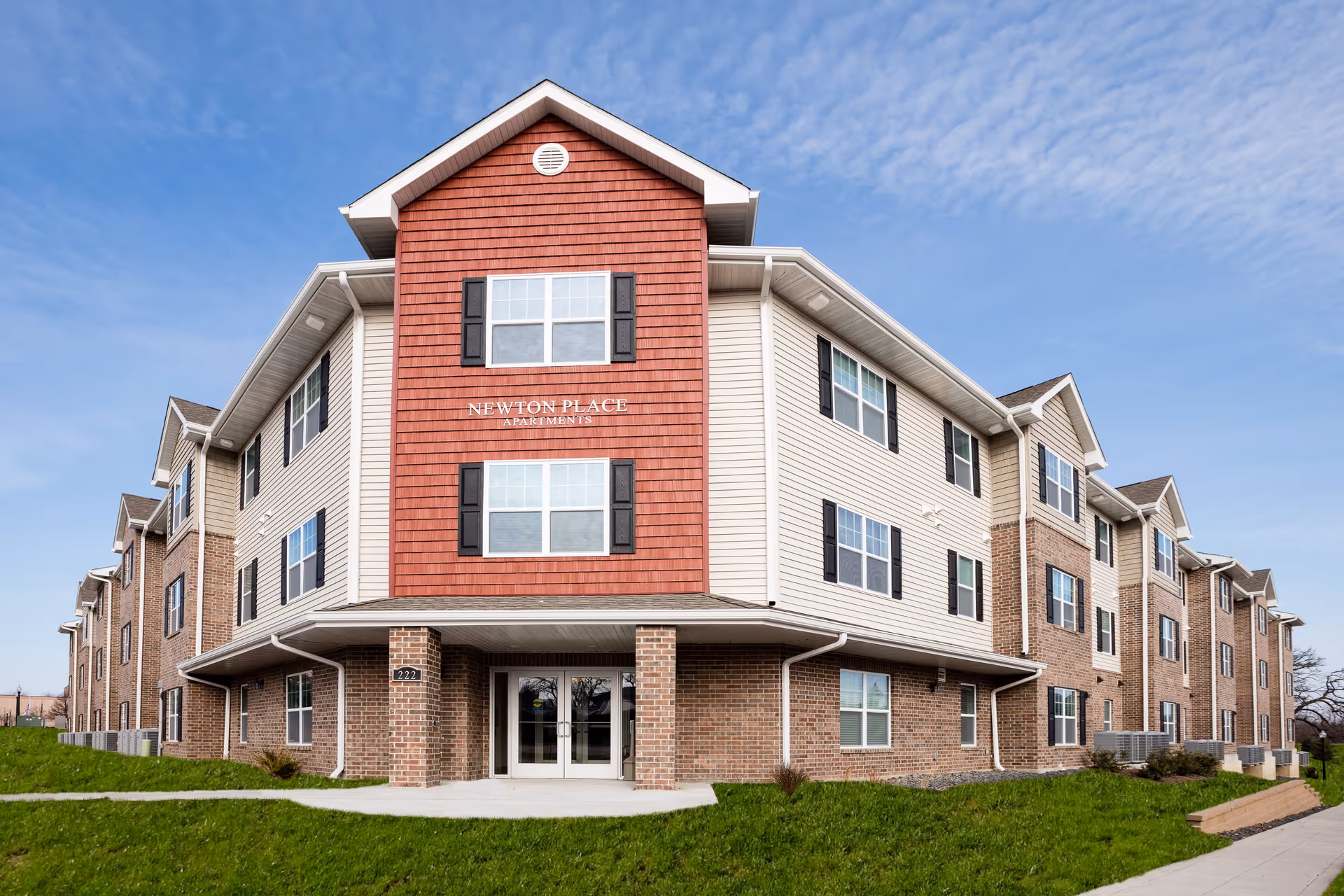 Exterior view of Newton Place Apartments, a three-story residential building with a combination of brick and beige siding, black window shutters, and a red gable section displaying the building name. The building is surrounded by green grass and a sidewalk under a partly cloudy blue sky.