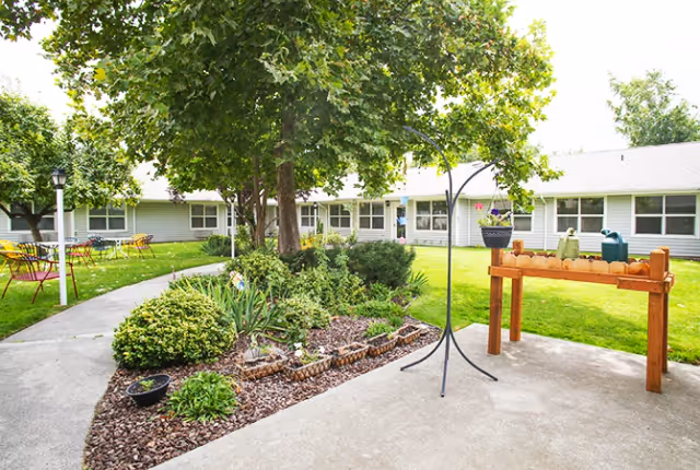 Outdoor courtyard area of a senior living facility with a concrete walkway, green grass, trees, shrubs, and a raised wooden planter. There are several chairs and tables arranged on the grass near the building, which has multiple windows and a light gray exterior.