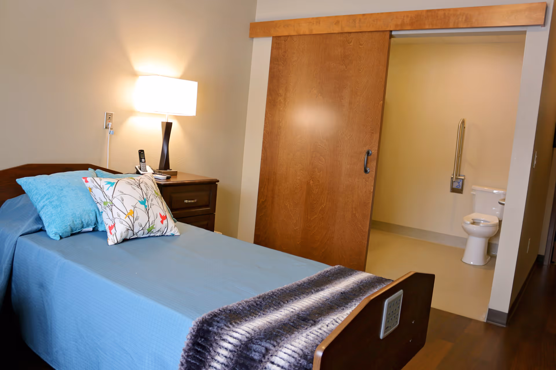A single bed with blue bedding and decorative pillows next to a wooden nightstand with a lamp and a phone. A sliding wooden door is partially open, revealing a bathroom with a toilet and a grab bar.